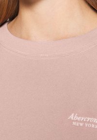 Jasnoróżowa bluza z okrągłym dekoltem, z prążkowaną fakturą, z subtelnym teksturowanym materiałem i haftowanym logo "Abercrombie New York" w białym kolorze.