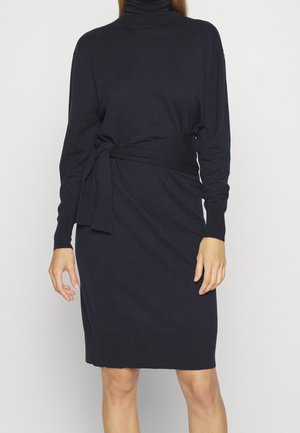 Mujer vestida con un vestido negro de manga larga y cuello alto, con un cinturón atado alrededor de la cintura, posando frente a un fondo liso.