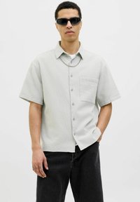 Camisa de manga corta, gris claro, de botón con una tela texturizada, un bolsillo en el pecho y botones azules, combinada con pantalones oscuros y gafas de sol.
