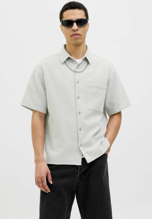 Camisa de manga corta, gris claro, de botón con una tela texturizada, un bolsillo en el pecho y botones azules, combinada con pantalones oscuros y gafas de sol.