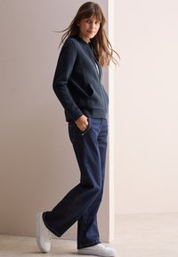 Giacca a zip blu marina con un motivo a spina di pesce testurizzato, abbinata a jeans in denim blue scuro a gamba larga e sneakers bianche.