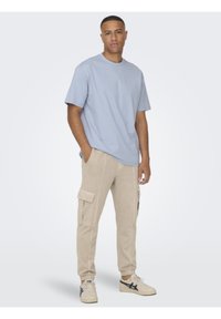 Hellblaues Baumwoll-T-Shirt mit rundem Ausschnitt, kombiniert mit beigen Baumwoll-Cargohosen mit Seitentaschen und gerippten Abschlüssen. Weiße Sneaker mit schwarzen Akzenten.