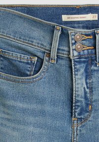 Vaqueros ajustados en denim azul medio con una textura desgastada. Presenta un cierre de botón, cinco bolsillos y costuras en naranja de contraste. Etiqueta de tamaño: 27.