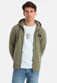 Sudadera con capucha en verde olivo con un acabado texturizado, que presenta dos bolsillos frontales y un pequeño parche de logo negro en el pecho. Lleva puesta sobre una camiseta blanca.