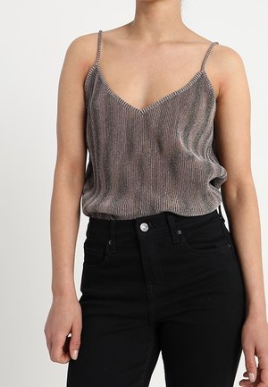 Person iført en metallisk, ribbet spaghetti-strop top stukket ned i sorte højtaljede jeans, stående mod en ensfarvet lys baggrund.