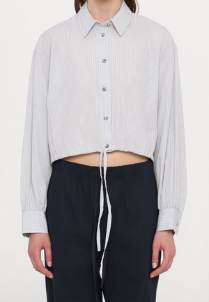 Femme portant une chemise courte rayée légère avec des manches longues et un pantalon ample foncé, montrant un cordon à la taille.