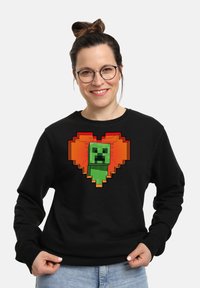 Felpa nera con un personaggio verde pixelato al centro di un design a forma di cuore con accenti arancioni. Materiale in cotone, vestibilità casual.