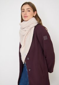 Žena nosící velký béžový šátek a tmavě fialový kabát s knoflíky a modré džíny, stojící před jednoduchým světlým pozadím.