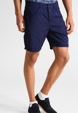 Mężczyzna ubrany w granatowe szorty z podwiniętymi brzegami, białe skarpetki do kostek i czarne trampki, stojący z nogami lekko rozstawionymi.