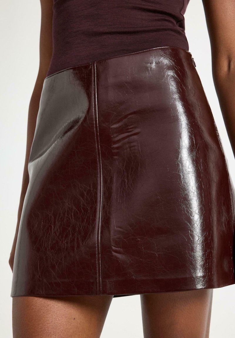 Mini gonna in pelle lucida marrone scuro con cucitura visibile e leggere pieghe, indossata con un top fitted viola scuro.