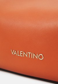 Sac en cuir orange avec une surface texturée et une inscription dorée "VALENTINO" en bas. Présente un design minimaliste.