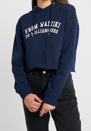 Femme portant un sweat à capuche court bleu marine avec l'inscription « From Waikiki to Williamsburg » et un jean noir taille haute, les mains jointes de manière détendue devant.