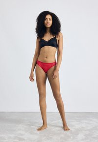 Black lace bralette with scalloped edges paired with red high-cut panties featuring lace trim. Model stands barefoot on a light gray surface.