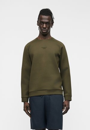 Mees punutud juustega, kes kannab oliivrohelise tooniga dressipluusi minimaalse logoga ja tumesiniseid lühikesi pükse halli tausta ees.