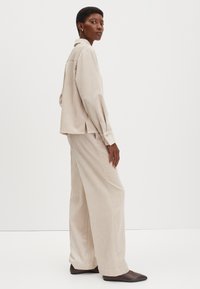 Beige Corduroy-Hemd mit Kragen, kombiniert mit passenden weit geschnittenen Hosen. Das Design bietet vertikale Streifen und seitliche Schlitze, getragen mit dunklen spitzen Schuhen.