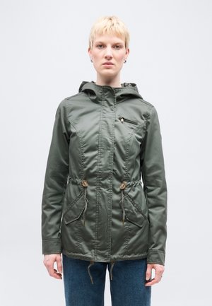 Person med kort blondt hår som har på seg en grønn hettejakke med snor og glidelåslommer, stående foran en ensfarget hvit bakgrunn.