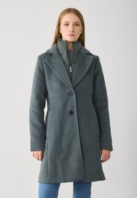 Cappotto lungo verde in lana con rever a punta, due tasche frontali e tre bottoni. Giacca interna leggera con colletto alto e cerniera.