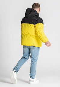 Puffjakke med sort hætte og øverste sektion, lys gul krop, blank finish og lange ærmer, båret over lyseblå jeans og hvide sneakers.