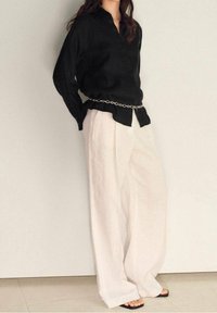 Chemise noire à manches longues avec col, associée à un pantalon large en lin blanc et une ceinture en lien circulaire argenté. Des sandales plates noires complètent le look.