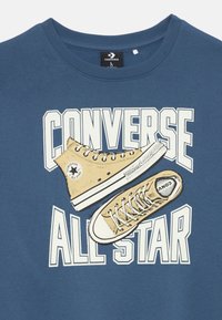 Camisa azul con el gran texto blanco "Converse All Star" y una ilustración de unas zapatillas Converse de caña alta en color beige sobre el texto.
