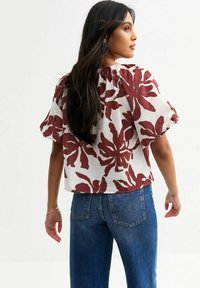 Blouse florale blanche avec des motifs de feuilles bordeaux, décolleté froncé, manches courtes bouffantes, associée à un jean en denim bleu.