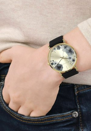 Handgelenk mit schwarzem Armband, Uhr mit goldenem Gehäuse und Zifferblatt mit Blumenmuster, getragen über dem Ärmel eines beigen Pullovers und blauen Jeans.