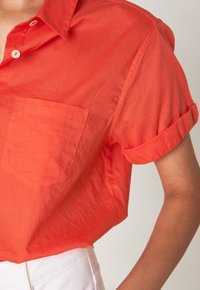 Camicia arancione a maniche corte con bottoni, realizzata in tessuto leggero, caratterizzata da una tasca applicata e polsini arrotolati, presentata su uno sfondo neutro.