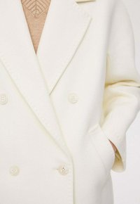 Dubbelzijdige jas van zacht witte stof, met een notch-kraag, twee voorzakken en lichtgekleurde knopen. De basislaag is beige.