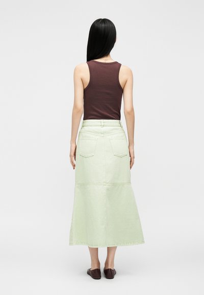 Femme aux longs cheveux noirs raides, portant un top marron sans manches, une jupe midi vert clair avec des poches arrière et des ballerines marron, debout de dos.