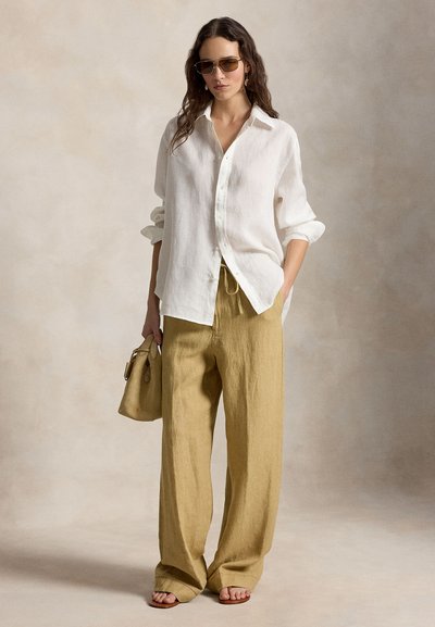Γυναίκα που φοράει υπερμεγέθη λευκό πουκάμισο με κουμπιά, χαλαρό μπεζ παντελόνι, γυαλιά ηλίου, κρατώντας μπεζ τσάντα, στέκεται μπροστά σε ουδέτερο φόντο.