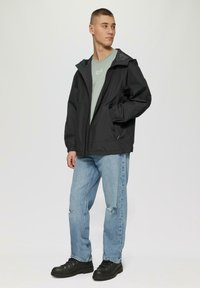 Zwarte lichtgewicht jas met capuchon, rits aan de voorkant, elastische manchetten, gedragen over een lichtgroen shirt, gecombineerd met blauwe jeans en zwarte laarzen.