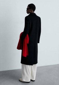 Zwarte wollen jas met een getailleerd ontwerp. Een felrood sjaal wordt in één hand vastgehouden. Crème brede broek en lichte schoenen maken de look compleet.