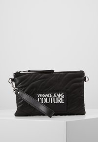 Svart quiltad clutchväska med dragkedjestängning, märkespatch och avtagbar rem. Tillverkad av glansigt material, med slät textur och lätt lyster.