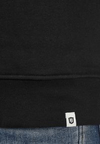 Czarna bluza z prążkowanym dołem i teksturowanym materiałem; posiada małą białą metkę z logo. Nosi się ją na niebieskim materiale dżinsowym.