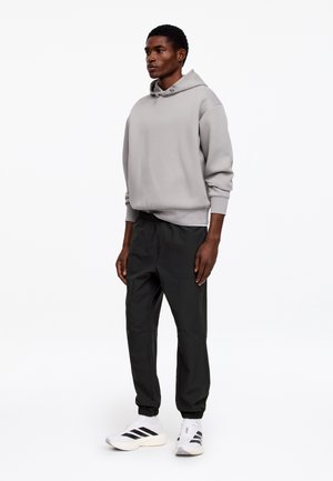 Mężczyzna stoi, ubrany w jasnoszary kaptur, czarne spodnie dresowe oraz białe trampki z czarnymi paskami, na jednolitym białym tle.