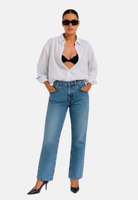Jeans blu a gamba dritta abbinati a una camicia over size bianca a righe, un reggiseno in pizzo nero e tacchi alti neri. Gli occhiali da sole completano il look.