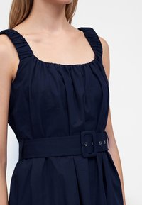 Femme portant une robe bleu marine sans manches avec des bretelles froncées et une large ceinture assortie avec une boucle rectangulaire.