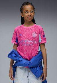 Rosa Manchester City-tröja med ett texturerat mönster, med blå accenter och logotyp. Buren med en blå pufferjacka och denimshorts.