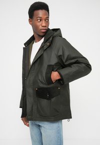 Jachetă verde închis cu glugă, având un buzunar texturat din catifea neagră, nasturi capse și croială lejeră. Buzunare laterale și frontale vizibile.