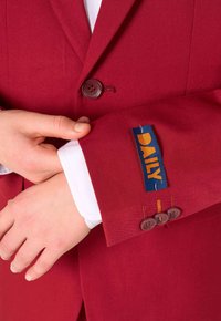 Sarkans žakets ar divām brūnām pogām, baltu krekla piedurkni un zilu uzlīmi ar tekstu "DAILY", kas iestrādāta piedurknē. Strukturēta auduma.