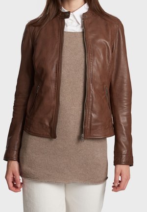 Femme portant une veste en cuir marron par-dessus un pull beige en maille et une chemise blanche à col, associée à un pantalon blanc, sur un fond uni.