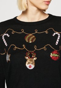 Schwarzer Pullover mit bunten, glitzernden Weihnachtsmotiven: Zuckerstangen, ein Rentier, Christbaumkugeln und ein silbernes Blatt, alle in Gold umrandet.