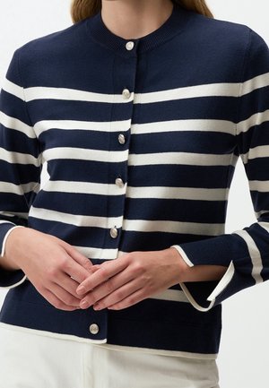 Kvinde iført en marineblå og hvid vandretstribet cardigan med sølvknapper, kombineret med hvide bukser, hænderne foldet foran livet.