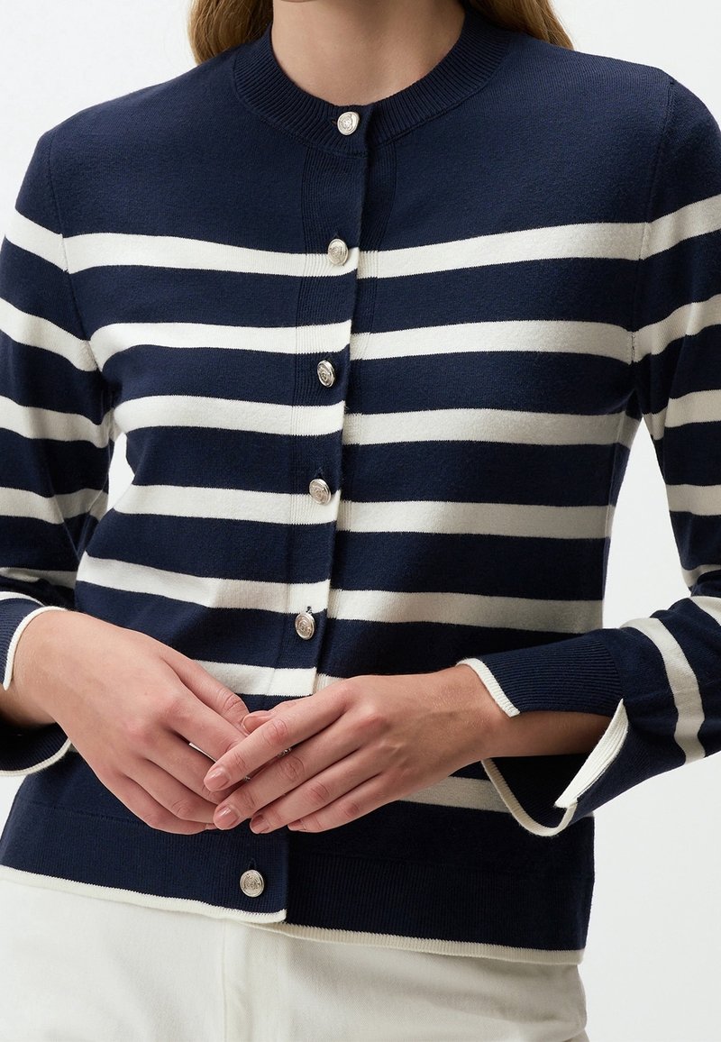 Femme portant un cardigan à rayures horizontales bleu marine et blanc avec des boutons argentés, associé à un pantalon blanc, mains jointes à la taille.