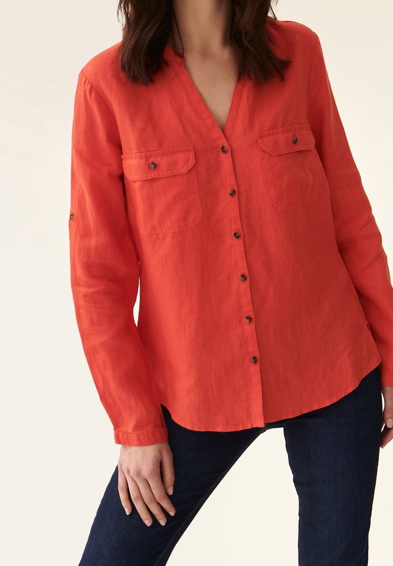Femme portant une chemise rouge à manches longues boutonnée avec deux poches devant et un pantalon bleu foncé sur un fond uni.