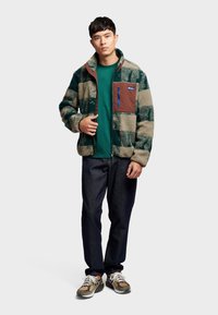 Fleece jacket in green and beige check pattern, featuring a brown chest pocket with blue zipper. Paired with a green t-shirt and dark jeans.