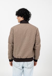 Mann mit lockigem, dunklem Haar, der eine braune Wolljacke mit dunklen gerippten Bündchen und Kragen sowie hellblauen Jeans trägt und mit dem Rücken zur Kamera steht.