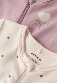Deux bodies pour bébés : l'un est rose clair avec un motif de cœur en forme de fleur, et l'autre est crème avec de petits cœurs gris. En matière de coton doux.