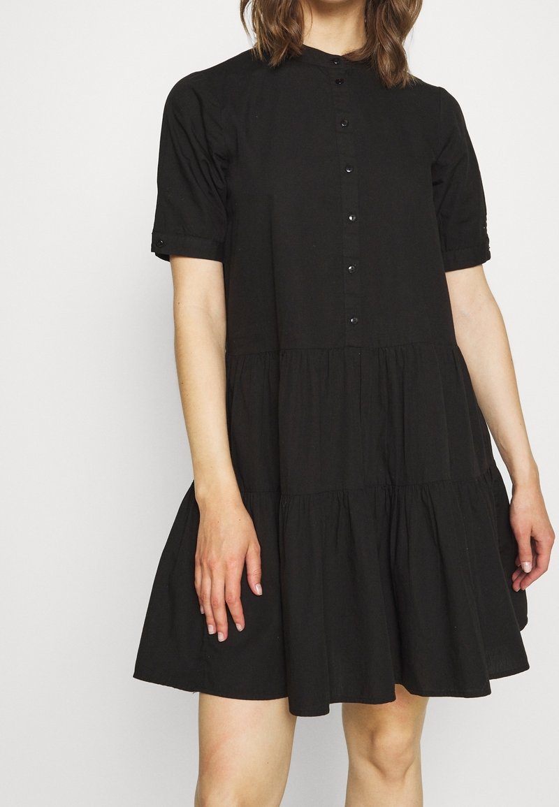 Femme portant une robe noire courte à boutons avec manches courtes et jupe froncée, debout devant un fond clair uni.