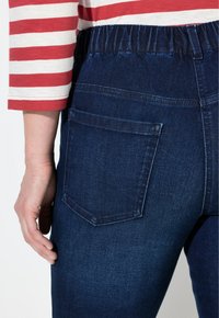 Záběr na osobu v tmavě modrých elastických džínách s pasem a v červeno-bílém pruhovaném tričku s dlouhým rukávem, s rukou uvolněně položenou na boku.